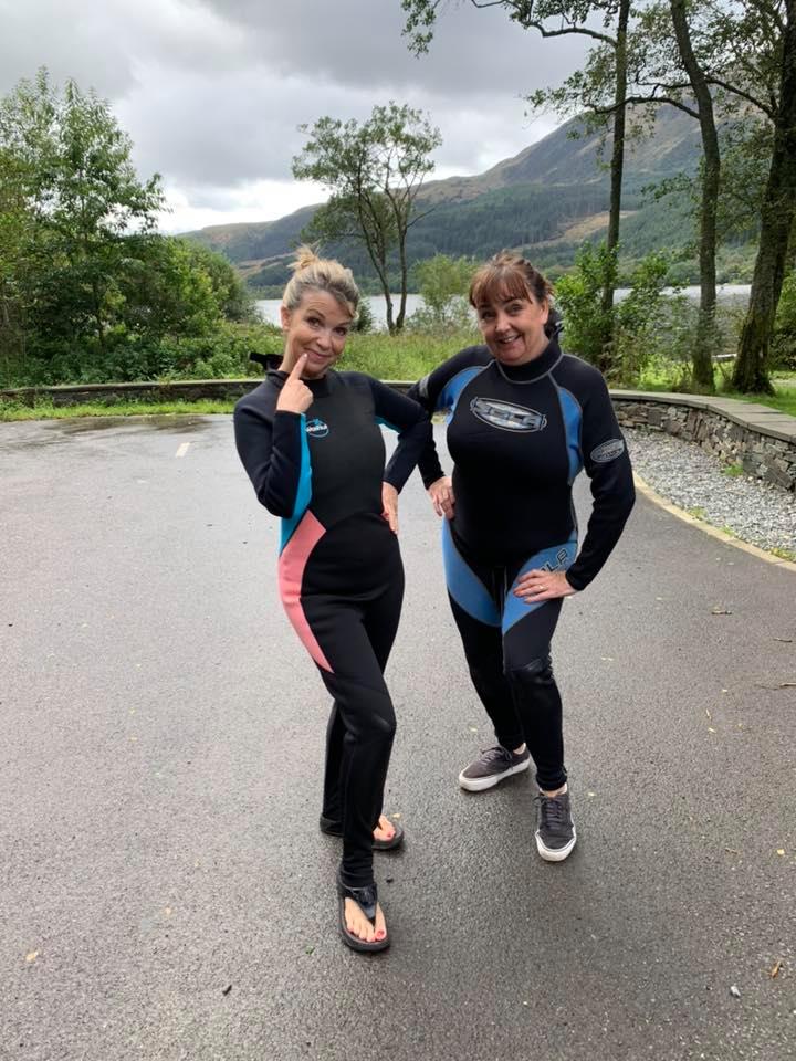 Two female Wild Swimmers in wetsuits, with lake and mountains behind them 